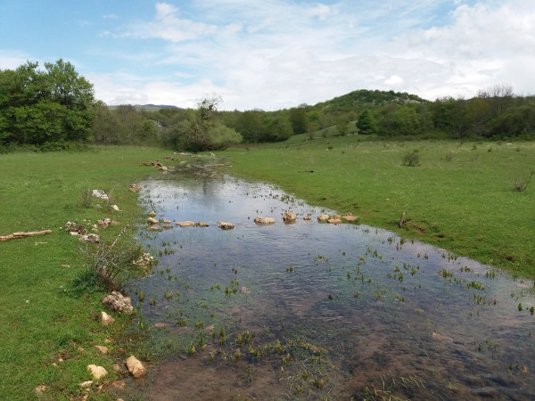 Prijelaz preko Rakića potoka koji se ulijeva u Jadovu. Iza desno brdo Stražbenica, jedno od najvažnijih arheoloških nalazišta u Lici iz doba Japoda.